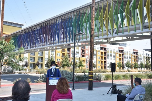 The image displays a person at a podium speaking to an audience for the Montague Pedestrian Overcrossing opening. The elevated pedestrian overcrossing is displayed behind the man at the podium.